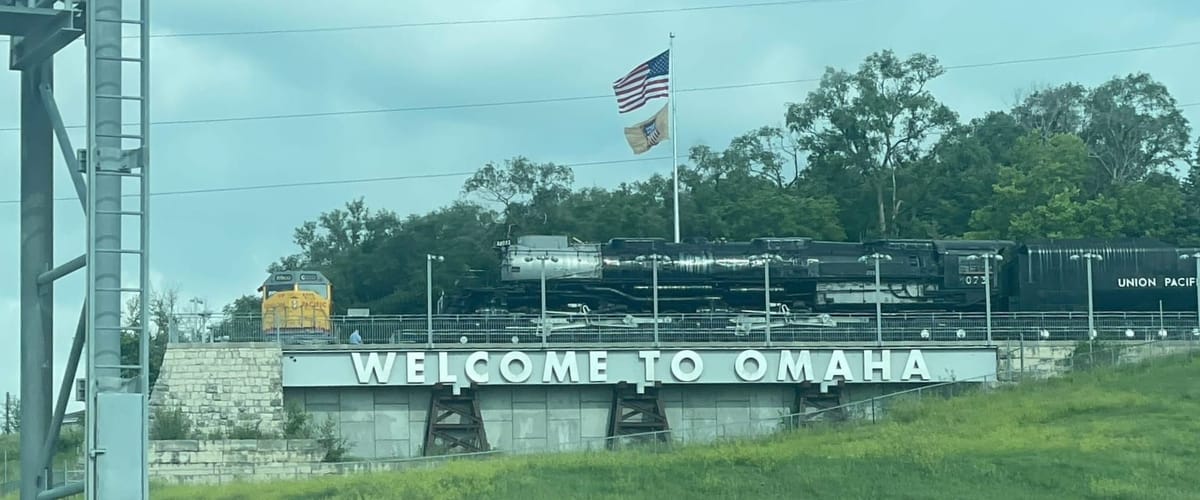 A black and silver steam locomotive facing left to right, on a bluff above a sign that says "Welcome to Omaha". Further left is a yellow diesel electric locomotive facing the camera